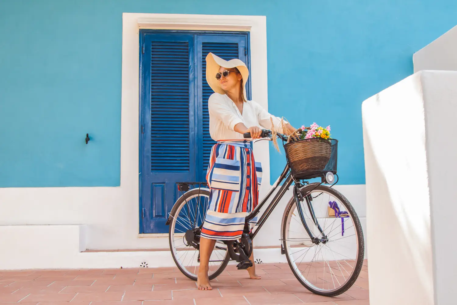 Ferry to Ponza - Beautiful young girl with bicycle in front of a blue house wearing colorful fashion dress with large hat in Sunny summer in Ponza island, Italy.