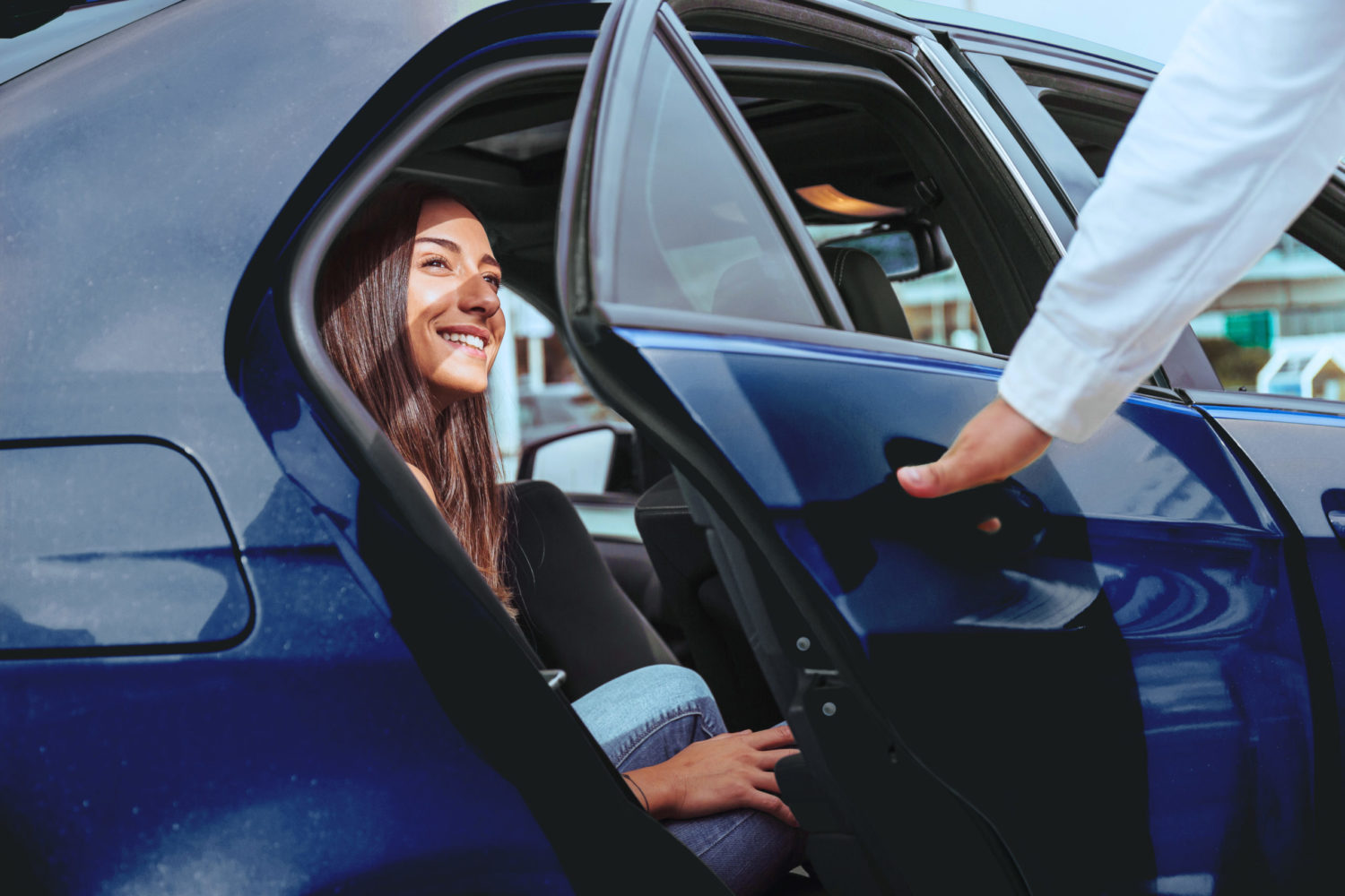 The image shows a young woman sitting in the back seat of a blue private transport vehicle. Her driver closes the door.