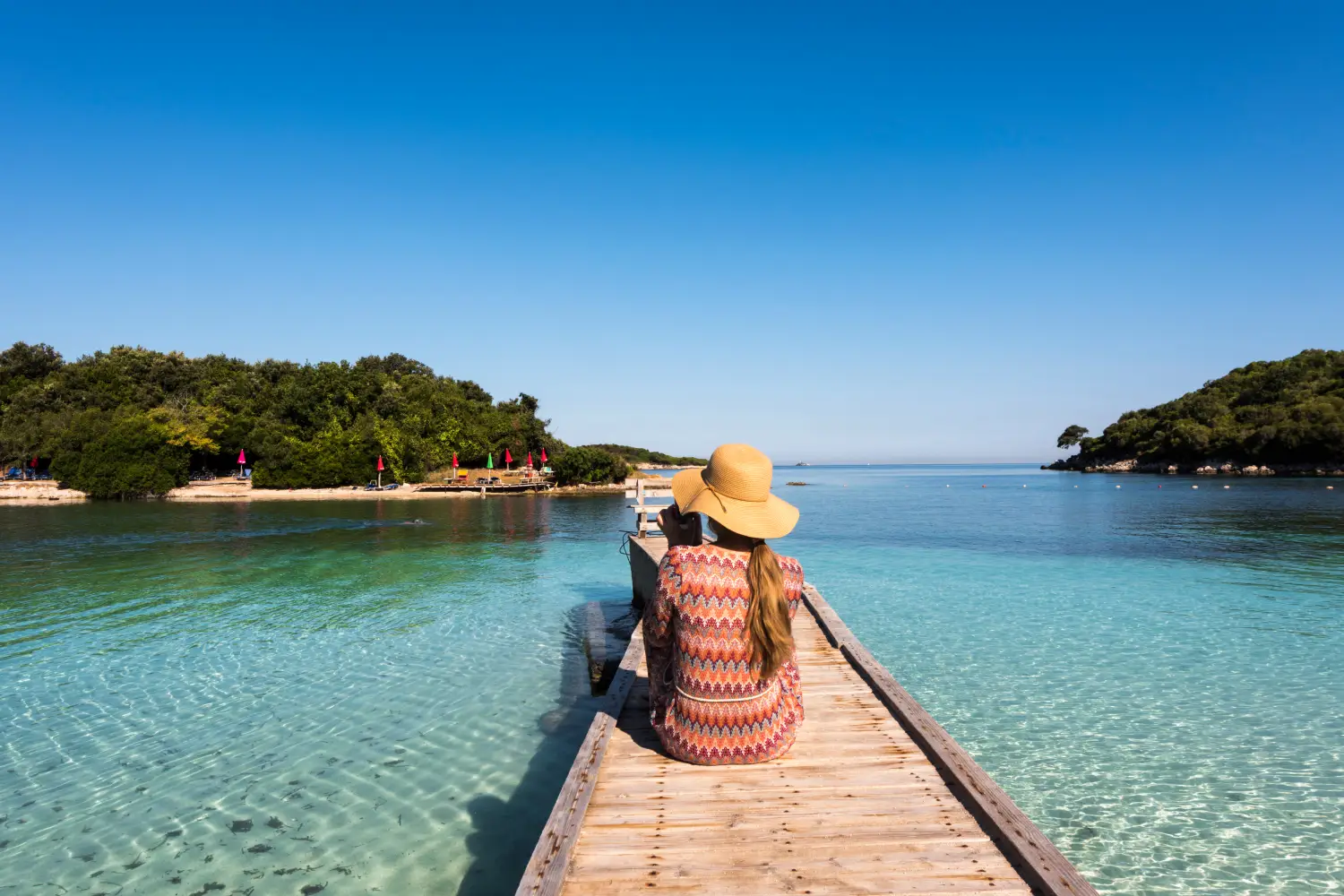 Ferry to Albania - Ksamil Beach - Woman with straw hat sitting on a wooden walkway, Ksamil, Albania.