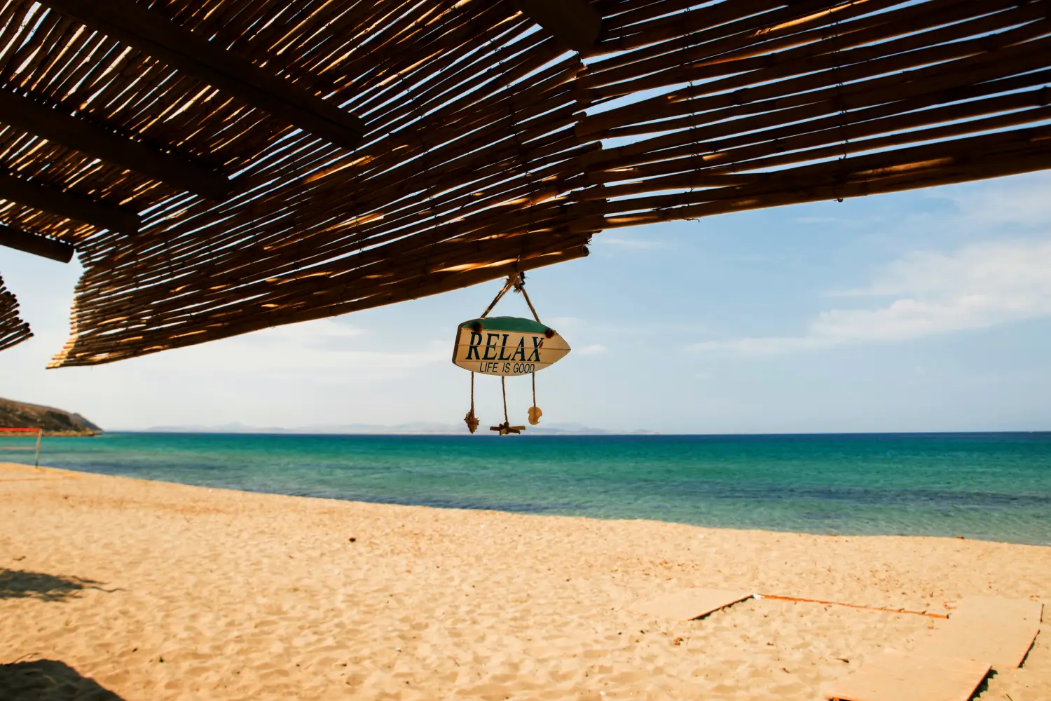 Ferry to Limnos - Empty sand beach and wicker sunshade with hanging text on small surfboard life is good, turquise blue seawater, island Limnos, Greece.